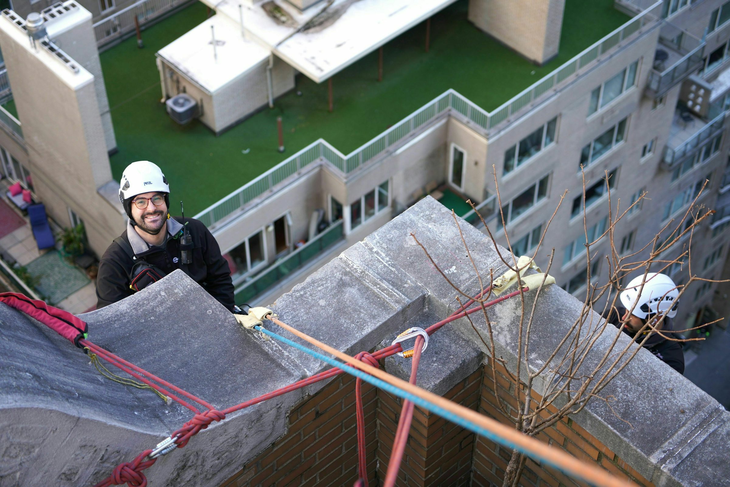 Two rope access workers wearing safety helmets and harnesses are preparing for a task on the edge of a tall building. One worker is smiling at the camera, while the other is partially visible, focused on securing the ropes. The background features modern residential buildings with green rooftop terraces.