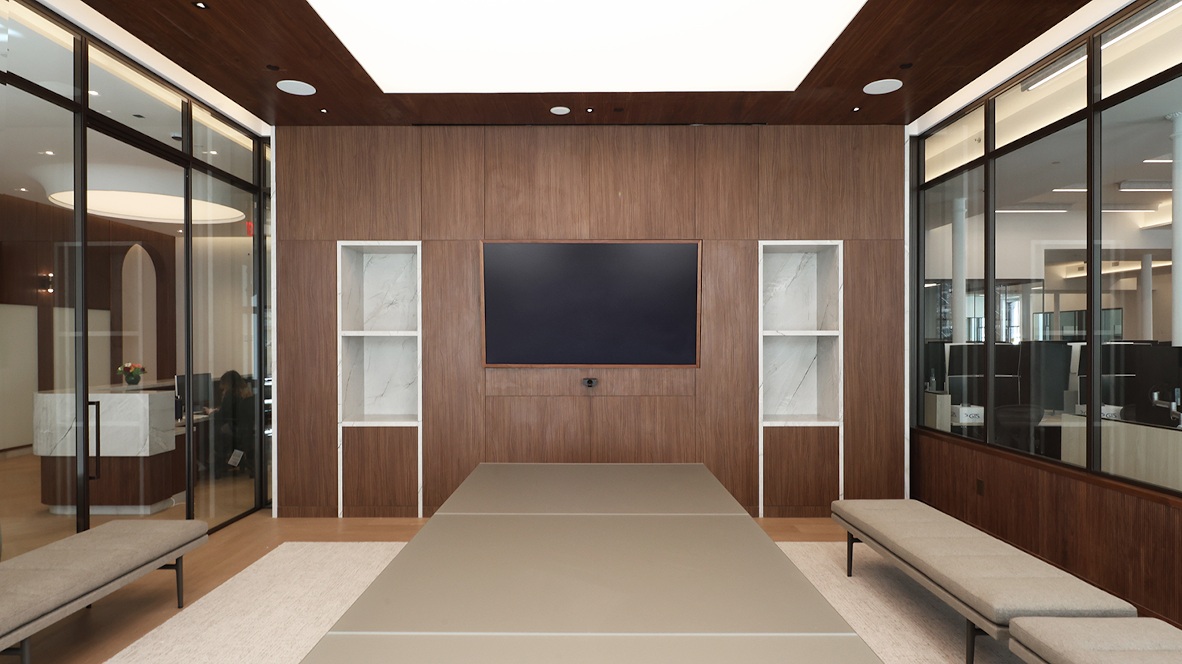 Contemporary conference room with a wall-mounted screen, wood paneling, built-in marble shelving, glass walls, and a long meeting table with modern seating.