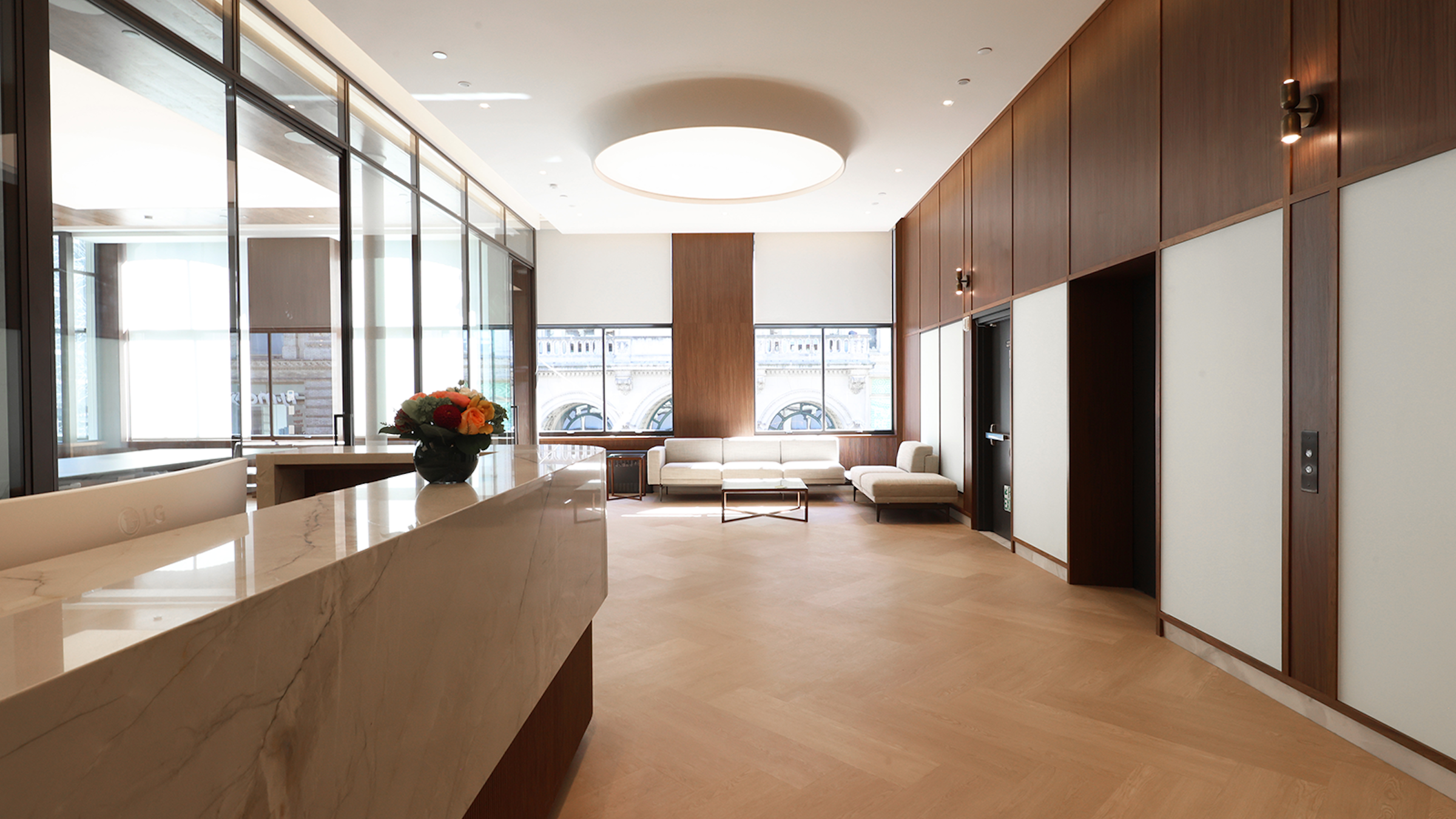 Modern office lobby with a marble reception desk, two people working, floor-to-ceiling windows, wood-paneled walls, neutral-toned furniture, and natural light.