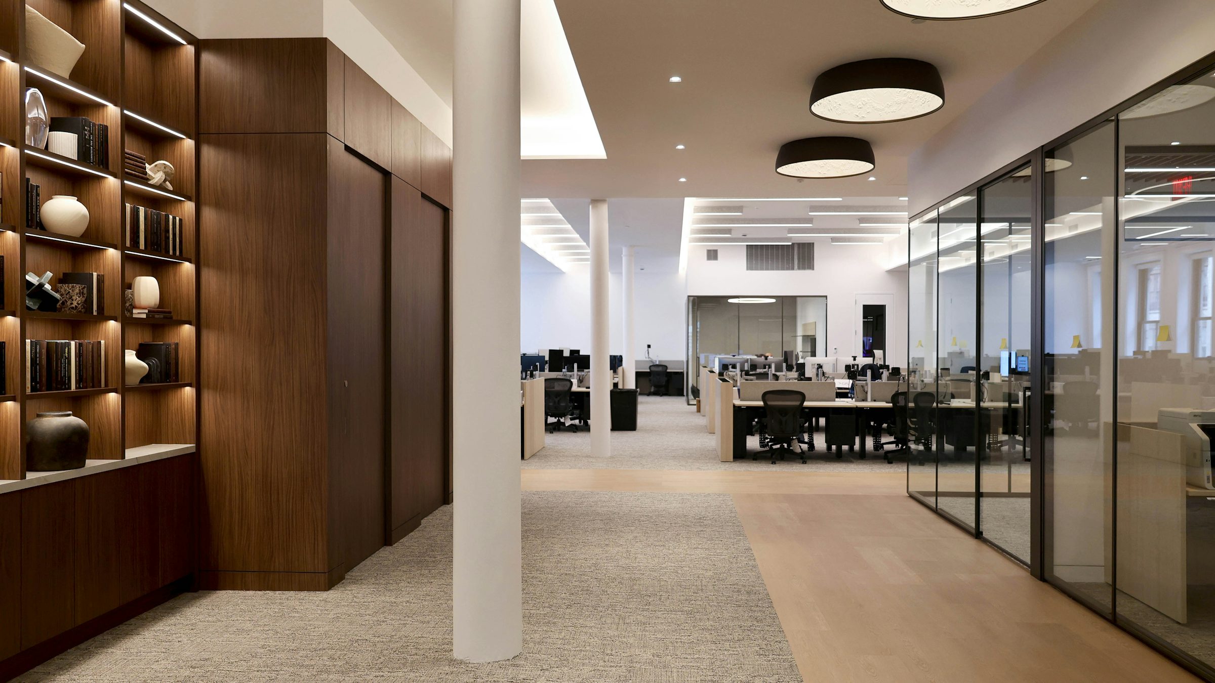 Modern open-plan office with glass-walled conference rooms, wood shelving with decorative objects and books, built-in lighting, and rows of minimalist workstations.